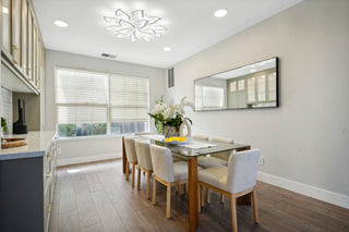 Room_with_a_glass_table_six_upholstered_chairs_a_large_wall_mirror_wooden_floor_and_a_floral_centerpiece_under_a_decorative_ceiling_light.