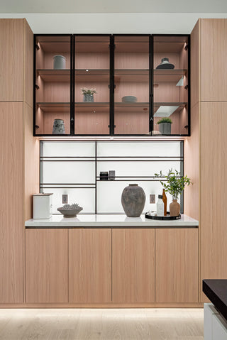 Sleek modern kitchen with light wood cabinetry, open shelves displaying decor, and a white countertop holding vases and plants, conveying a minimalist vibe.