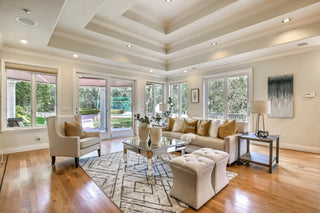 Spacious living room with large windows and wooden frames overlooking a lush garden. Cozy beige sofa with patterned cushions, a rustic wooden coffee table, and modern kitchen in background. Bright .