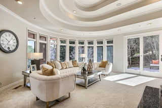 Spacious living room with large windows and wooden frames overlooking a lush garden. Cozy beige sofa with patterned cushions, a rustic wooden coffee table, and modern kitchen in background. Bright and .