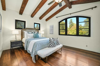 Spacious living room with large windows and wooden frames overlooking a lush garden. Cozy beige sofa with patterned cushions, a rustic wooden coffee table, and modern kitchen in background. Bright and inviting .