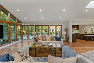 Spacious living room with large windows, wooden floors, and a cozy beige sofa. Blue rug and rustic coffee table enhance the modern, serene decor.