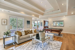 Spacious living room with larger windows and wood frames, white walls, and hardwood floors. Cozy beige sofas surround a rustic table. Airy and bright ambiance.
