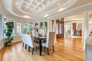Spacious living room with larger windows and wood frames, white walls, and hardwood floors. Cozy beige sofas surround a rustic table. Airyes and bright ambiance.