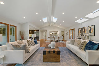 Spacious living room with two white sofas, adorned with patterned and blue cushions, facing a wooden coffee table. Skylights and modern art add elegance.