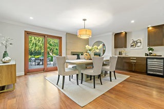 Warm and inviting dining room with a large wooden table, six beige chairs, and a cream rug. French doors open to a lush garden, enhancing a cozy, elegant ambiance.