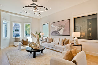  cozy living room features two white armchairs facing a low round table adorned with decor and a soft throw. Built-in shelves with warm lighting showcase vases and art, framing a black and white abstract painting above 