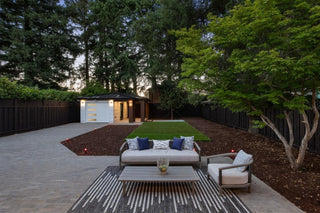 update house exterior at dusk with warm lights. Large glass doors open to reveal a stylish, inviting interior. Trees and a fence surround the patio.